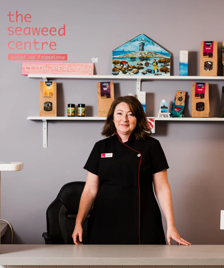 A woman in spa uniform standing at the front desk with seaweed products displayed on shelves behind her